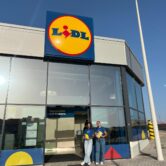 Dos personas sonrientes sosteniendo folletos frente a la tienda Lidl en El Médano, destacando la colaboración entre BlueFox y Lidl para acercar las marcas a los consumidores en el punto de venta.